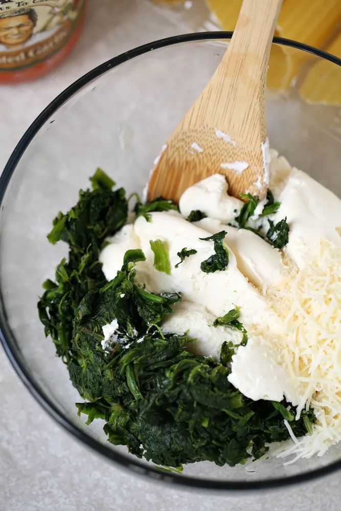 manicotti filling prepared with chopped spinach and ricotta cheese and shredded cheese in clear bowl with wooden spoon
