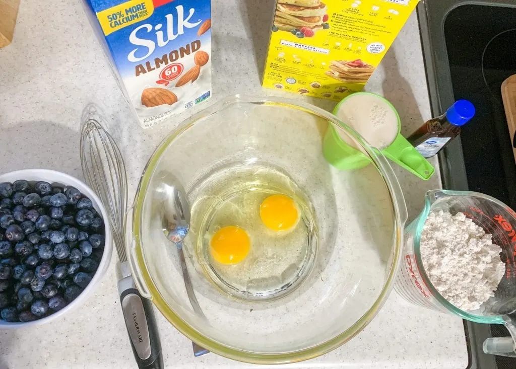 bowl of 2 eggs and blueberries, almond milk, whisk and pancake mix